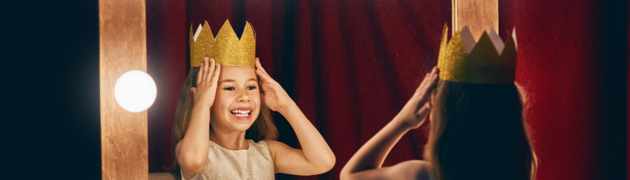 Lets_Make_Theatre_hero.png A girl looking in a dressing room mirror while straightening a paper crown.