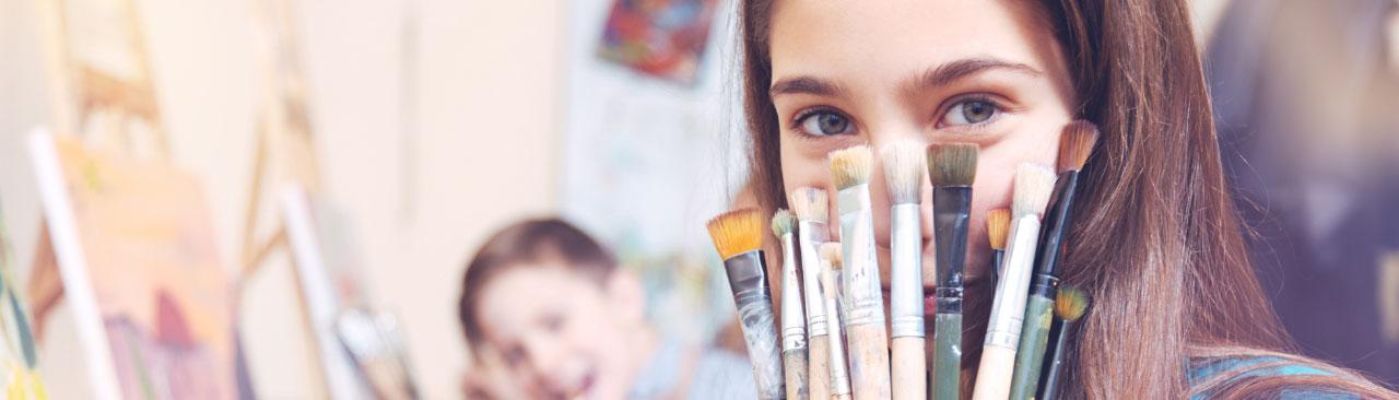 A teen looking over the top of a handful of paintbrushes